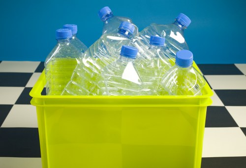A bright yellow plastic bin filled with several transparent plastic bottles, some of which are partially crushed or crumpled, each topped with blue caps. The bin is situated on a surface with a black and white checkered pattern, and the background features a plain blue wall. This scene likely depicts a collection of recyclable plastic bottles ready for disposal or collection, in a setting consistent with waste collection services such as those offered by Commercial Waste Gerrards Cross, serving the Gerrards Cross area. The bottles vary slightly in shape and size but predominantly appear lightweight and made of clear PET material, with smooth surfaces and subtle reflections of light. The arrangement shows the bottles leaning against each other, some standing upright while others are tilted or resting on their sides, giving a cluttered appearance. The image emphasizes the need for proper disposal of plastic waste within local waste management operations, situated in a clean, indoor environment suitable for rubbish collection or recycling activities.