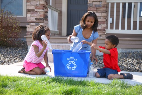 Three young children with varying skin tones are sitting on a white mat outdoors on a residential driveway, participating in recycling and rubbish collection activities. The central girl, wearing a light blue top, is actively placing clear plastic bottles into a large blue recycling bin labeled with a white recycling symbol and the words 'We Recycle.' To her left, a young girl in a pink and white striped dress is holding a clear plastic bottle near her mouth, preparing to place it into the bin. On the right, a boy dressed in a red shirt and dark shorts is also engaged in the process, holding a plastic bottle above the bin. The children are surrounded by a well-maintained garden with green grass, dark grey gravel, and some small plants. In the background, the entrance of a modern house with a dark grey door, stone and brick exterior walls, and white railings on the porch can be seen, suggesting a suburban setting in Gerrards Cross, Bucks. The scene is well-lit with natural daylight, emphasizing the environmental and rubbish management theme linked to Commercial Waste Gerrards Cross.